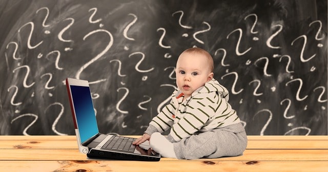 In front of a blackboard covered with question marks, a baby sits behind a laptop computer